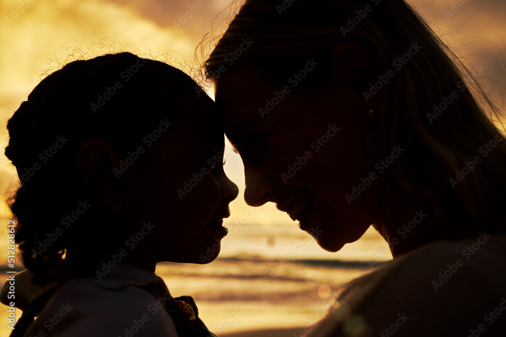 Closeup silhouette of a mother and daughter standing face to face on the beach at sunset. Backlit young woman and her girl child smiling and facing one another with the ocean in the background
