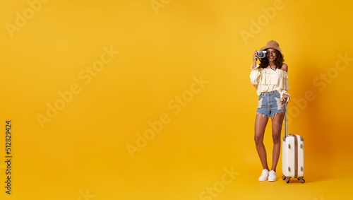 Young pretty african woman tourist smiling holding luggage with camera in hand isolated on yellow background.