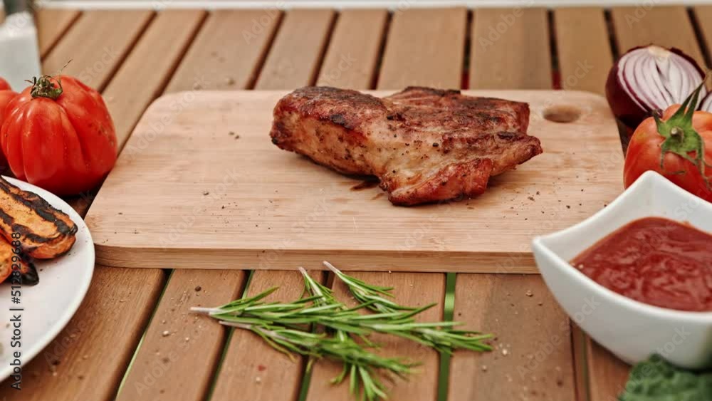 The hand of a cook with metal tongs puts a grilled pork steak on a piece of wood on a rustic table. The hand of a cook with metal tongs puts a grilled pork steak on a piece of wood on a rustic table.