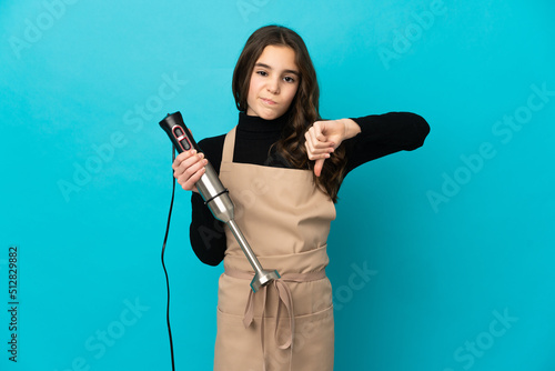 Little girl using hand blender isolated on blue background showing thumb down with negative expression
