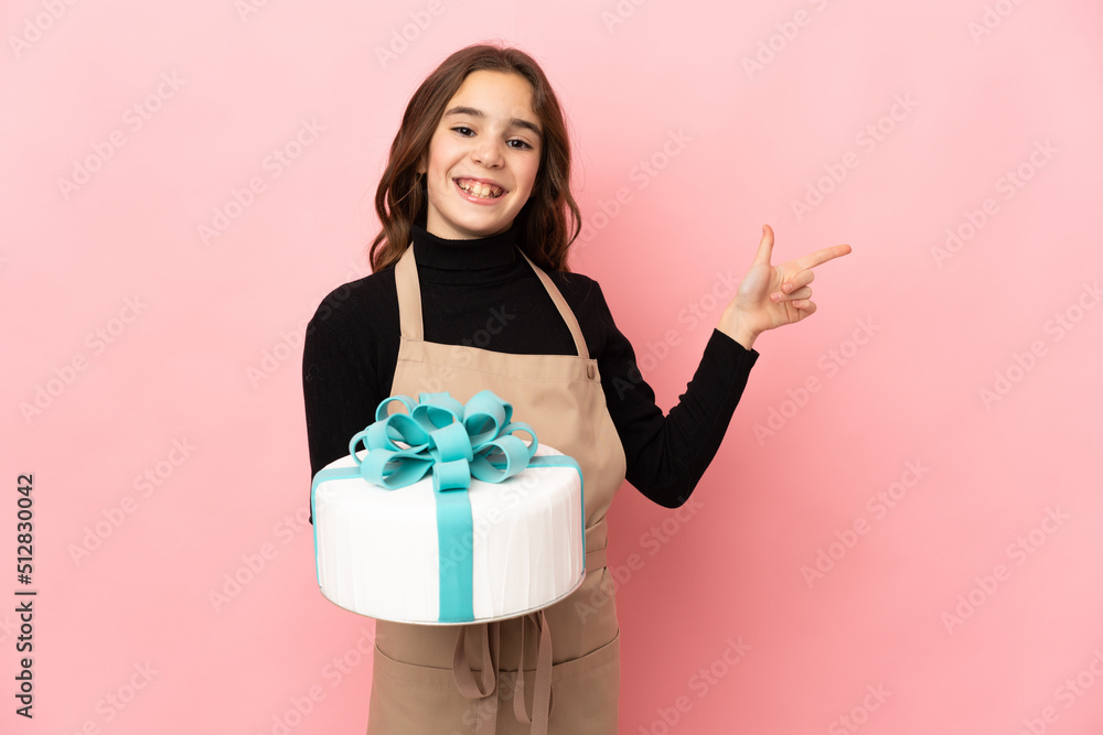 Little Pastry chef holding a big cake isolated on pink background pointing finger to the side Little Pastry chef holding a big cake isolated on pink background pointing finger to the side