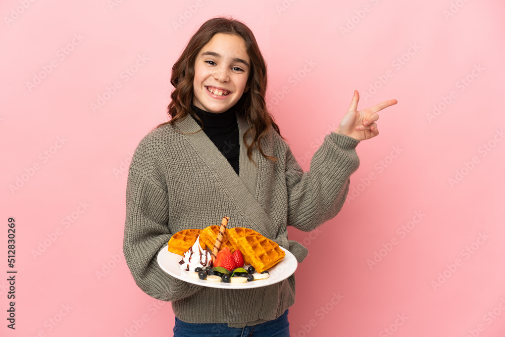Little girl holding waffles isolated on pink background pointing finger to the side Little girl holding waffles isolated on pink background pointing finger to the side