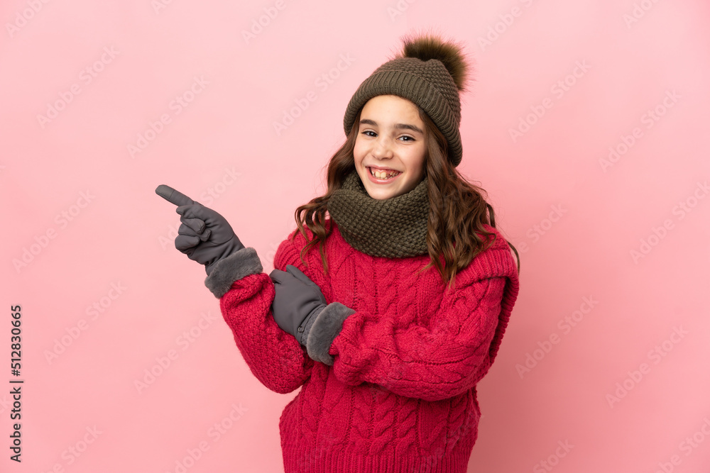 Little girl with winter hat isolated on pink background pointing finger to the side