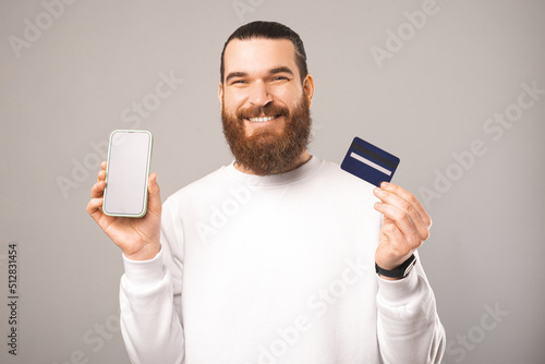Cheerful young bearded man is holding a phone and card showing screen.