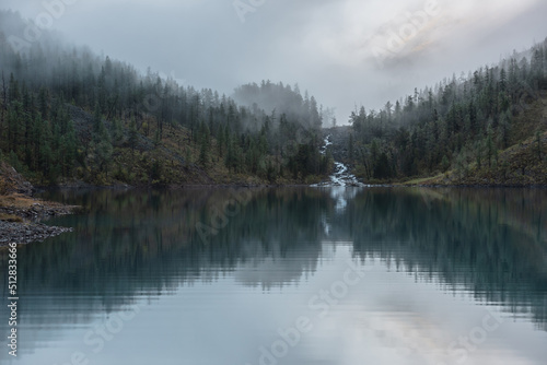 Mountain creek flows from forest hills into glacial lake in mysterious fog. Small river and coniferous trees reflected in calm alpine lake in early morning. Tranquil misty scenery with mountain lake. © Daniil