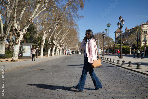 Young beautiful South American woman in a pink jacket, jeans, brown handbag and sunglasses, on vacation in Europe, crossing a city street. Concept vacation, travel, beauty, fashion. © Manuel