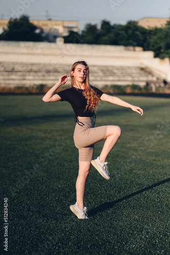 Woman in Black Top Exercising on Lawn Raising Her Knees Up. Sports Girl Illuminated by Golden Rays