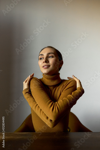 Self-esteem concept. Vertical image of arrogant woman hugging herself putting arms around her shoulders sitting at table against white wall. Cute office manager warming up during break