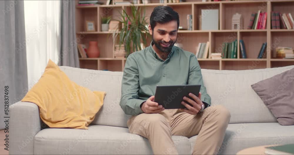 Handsome bearded arabian male with tablet speaking via camera sitting on sofa. Man discussing freelance online project on smartphone staying home during quarantine lockdown concept. Handsome bearded arabian male with tablet speaking via camera sitting on sofa. Man discussing freelance online project on smartphone staying home during quarantine lockdown concept.