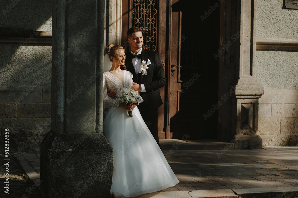 The bride and groom against the backdrop of beautiful architecture, gently embrace. The sun illuminates the tender faces of a beautiful couple. Gorgeous couple of Europeans. The bride and groom against the backdrop of beautiful architecture, gently embrace. The sun illuminates the tender faces of a beautiful couple. Gorgeous couple of Europeans.