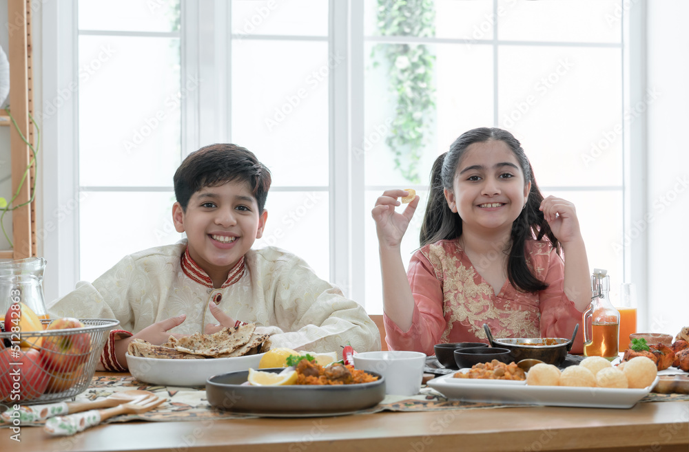 Happy Indian family enjoy eating food with hands, South Asian sibling, brother and sister smiling looking at camera, wearing traditional clothes, sitting at dining table at home together Happy Indian family enjoy eating food with hands, South Asian sibling, brother and sister smiling looking at camera, wearing traditional clothes, sitting at dining table at home together