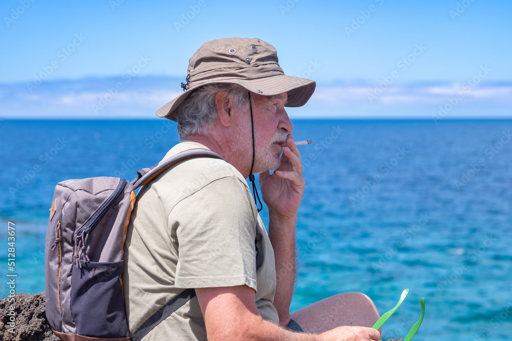 Relaxed senior man with hat and backpack sitting at sea smoking a cigarette, elderly grandfather looking away enjoying freedom and holidays