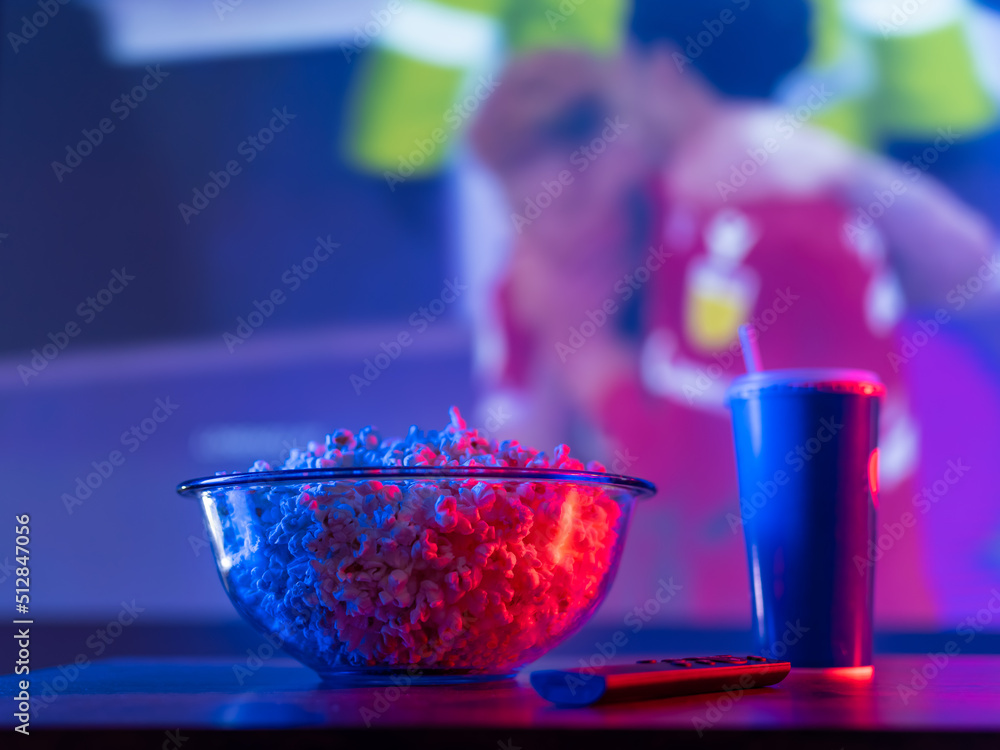 Fototapeta premium Basketball match on the TV screen. Popcorn, drink, TV remote on the table in front of the TV. Sports fan set. Watch sports programs in the comfort of your own home. Neon lighting.