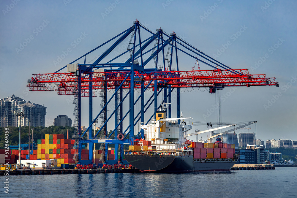 cargo ship moored for loading at container yard with container handling gantry crane or ship-to-shore crane unloading goods from deck to cargo port area, background cityscape with high-rise building. cargo ship moored for loading at container yard with container handling gantry crane or ship-to-shore crane unloading goods from deck to cargo port area, background cityscape with high-rise building.