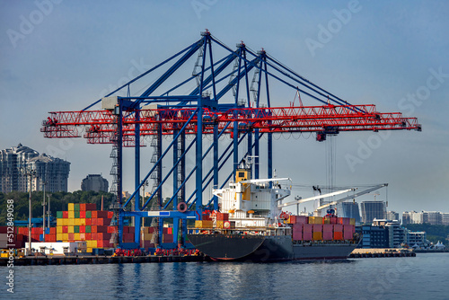 cargo ship moored for loading at container yard with container handling gantry crane or ship-to-shore crane unloading goods from deck to cargo port area, background cityscape with high-rise building.