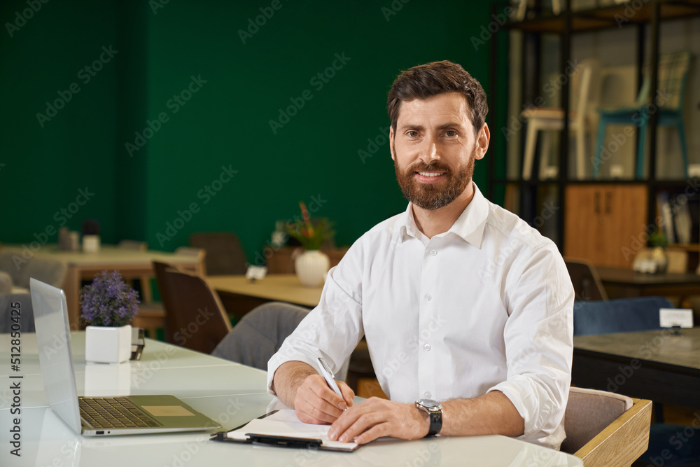 Elegant bearded man in shirt sitting at table with laptop, while working remotely at home. Portrait of handsome businessman looking at camera and smiling against green wall. Concept of workplace. Elegant bearded man in shirt sitting at table with laptop, while working remotely at home. Portrait of handsome businessman looking at camera and smiling against green wall. Concept of workplace.