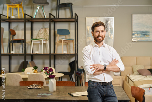 Confident shop owner in white shirt posing, with crossed arms in furniture store. Portrait of handsome businessman looking at camera, smiling, while leaning on table in showroom. Concept of business.