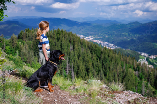 Girl with stands next to her dog of the Rottweiler breed on a peak with vegetation against the cloudy sky