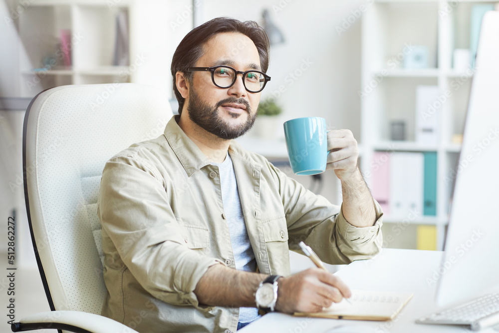 Portrait of handsome young Asian man in glasses sitting at table and making notes in sketchpad while enjoying morning coffee in office