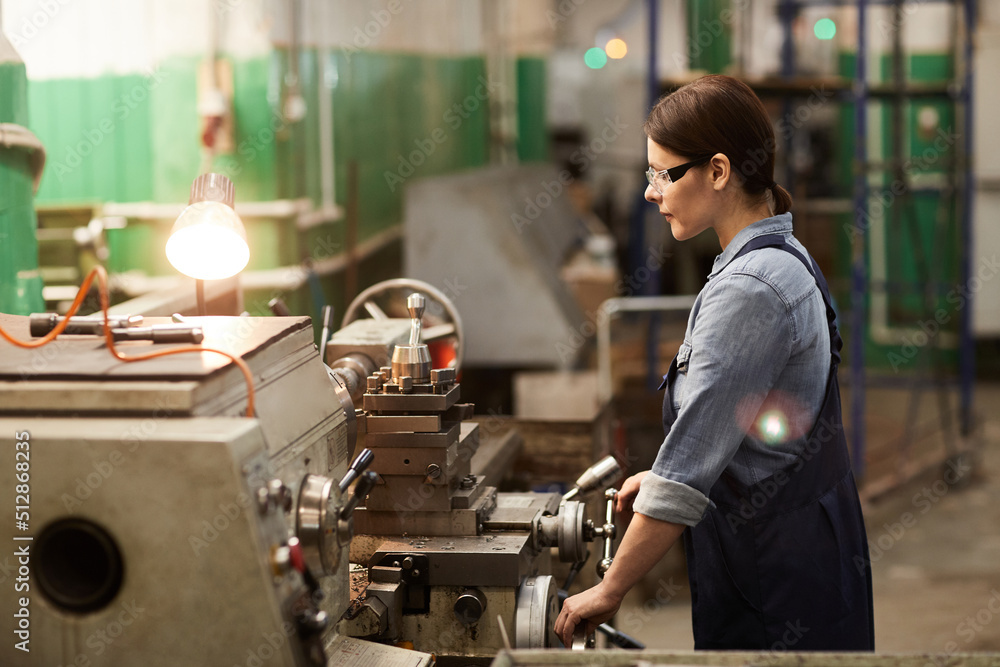 Concentrated middle-aged female industrial laborer in overalls susing manual lathe while processing metal detail Concentrated middle-aged female industrial laborer in overalls susing manual lathe while processing metal detail