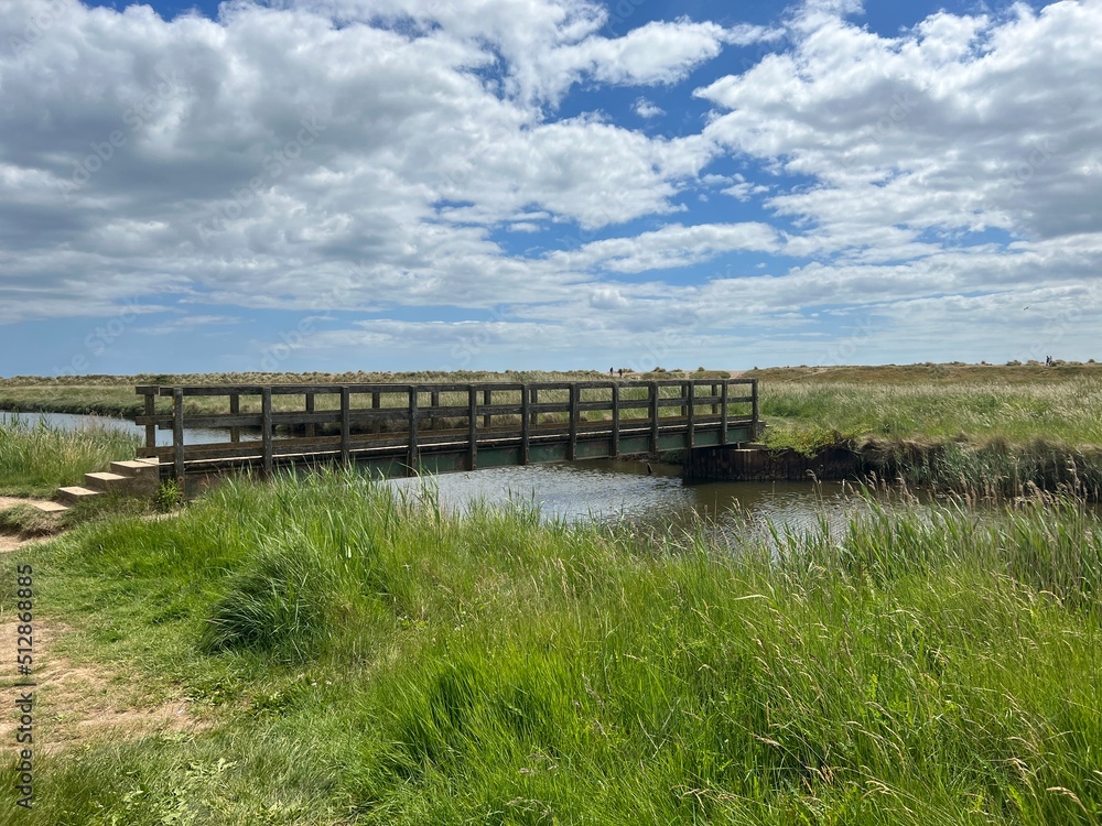 Beautiful landscape with wood bridge over waterway with reeds and sand path at nature reserve by the beach on East Anglia uk coastline at Walberswick Suffolk on Summer day with blue sky white cloud Beautiful landscape with wood bridge over waterway with reeds and sand path at nature reserve by the beach on East Anglia uk coastline at Walberswick Suffolk on Summer day with blue sky white cloud