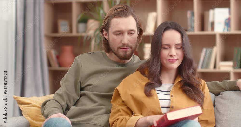 Close up on beautiful young caucasian couple man and woman sitting on sofa reading book together and looking into camera smiling. Happy people spending time indoors covid 19, quarantine concept. Close up on beautiful young caucasian couple man and woman sitting on sofa reading book together and looking into camera smiling. Happy people spending time indoors covid 19, quarantine concept.