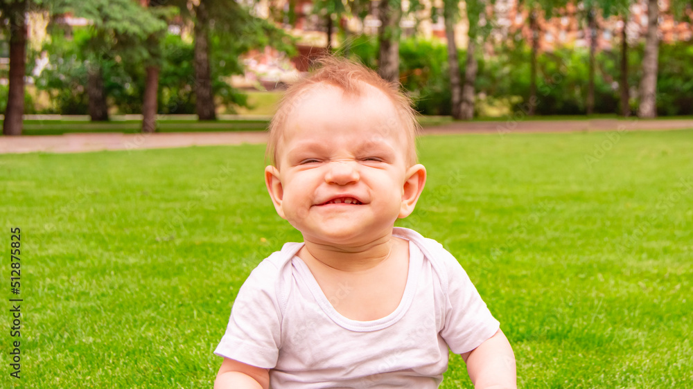 Cute laughing blonde baby girl 1 year old top having fun sitting in green grass outdoors. Childhood. Summer season. Looking at camera. Happy little child laughs nicker in meadow. Cute laughing blonde baby girl 1 year old top having fun sitting in green grass outdoors. Childhood. Summer season. Looking at camera. Happy little child laughs nicker in meadow.