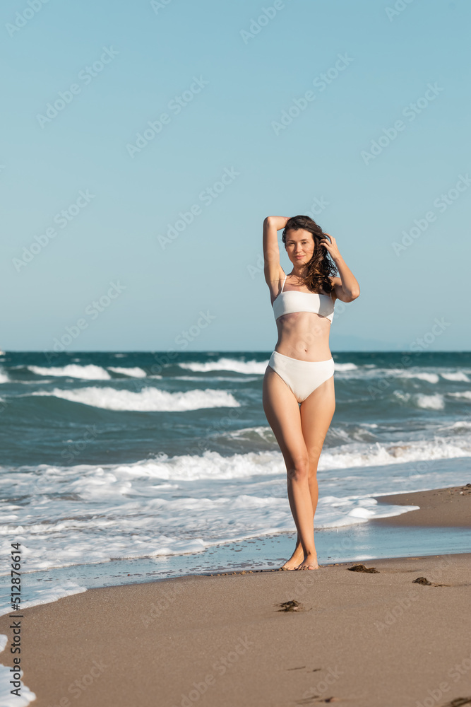 full length of smiling young woman in white swimsuit walking near ocean on beach. full length of smiling young woman in white swimsuit walking near ocean on beach.