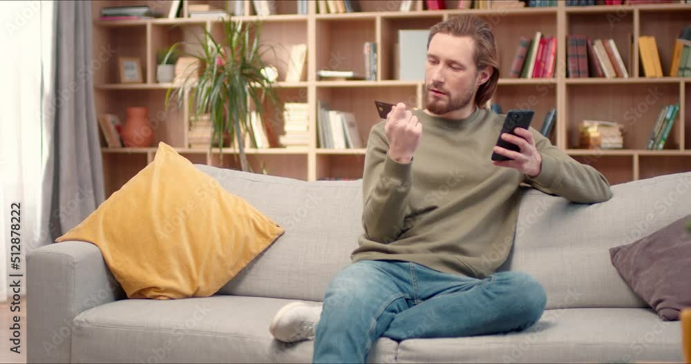 Happy caucasian man holding phone and credit card in hands sitting on sofa at home. Male shopper using instant easy mobile payments making purchase in online store. E-banking app service. Happy caucasian man holding phone and credit card in hands sitting on sofa at home. Male shopper using instant easy mobile payments making purchase in online store. E-banking app service.