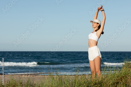 young sportive woman in shorts and sports bra stretching near blue sea.