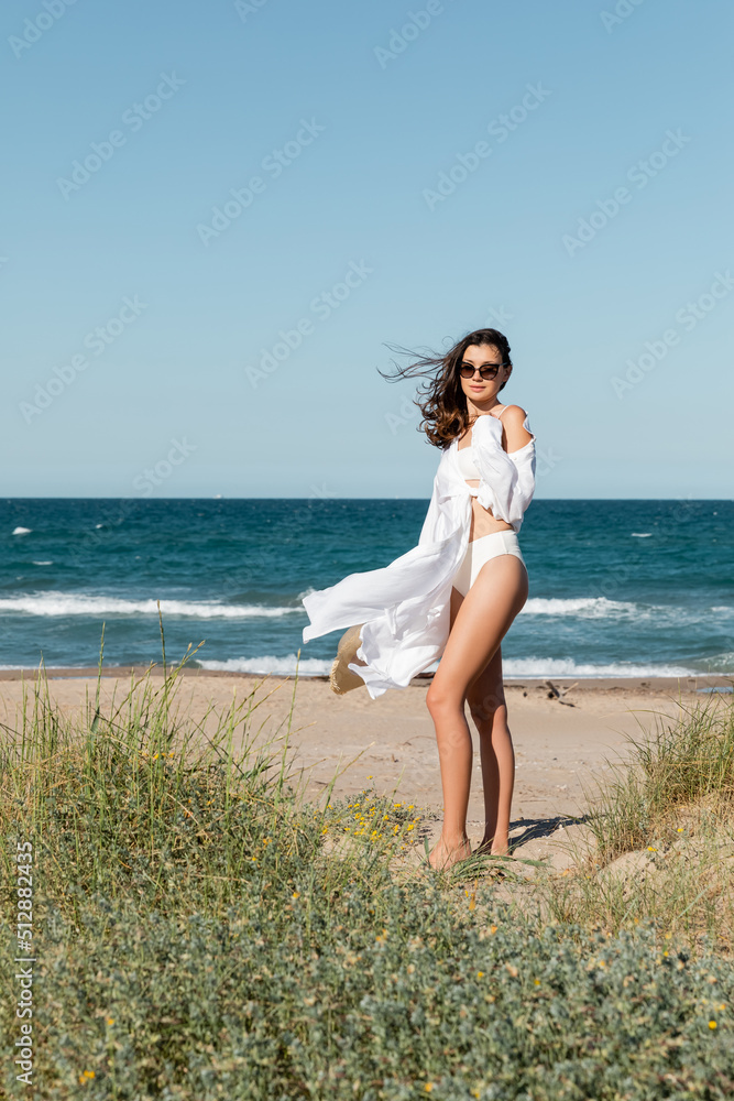 full length of brunette young woman in sunglasses and white shirt holding straw hat on sandy beach. full length of brunette young woman in sunglasses and white shirt holding straw hat on sandy beach.