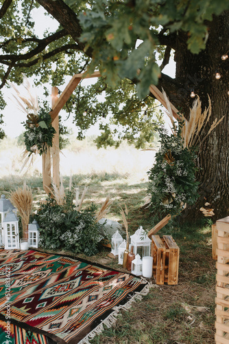 Wedding ceremoby decorations and elements in boho style. Ceremony near the ancient oak. Decorating with monstera leaves with candlesticks and wooden arch. Boho style rugs. With beautiful brown chairs.