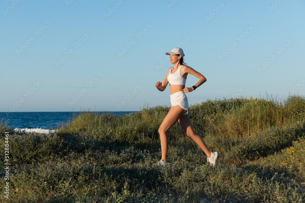 sportive woman in baseball cap and wireless earphone running on grass near sea. sportive woman in baseball cap and wireless earphone running on grass near sea.
