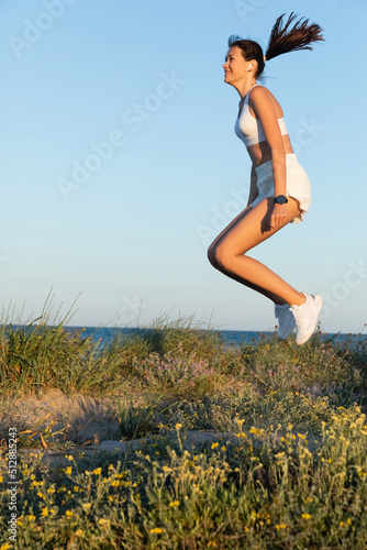 joyful and fit woman in sportive shorts and wireless earphone jumping near blue sea.