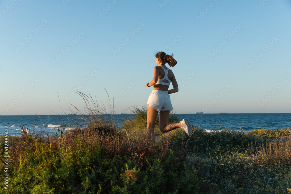 brunette woman in shorts and wireless earphone jogging on grass near sea shore.