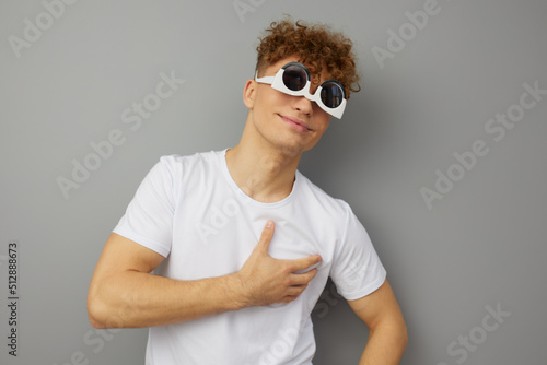 a playful young man stands in a casual T-shirt with funny glasses on his face and holding his hand on his heart standing on a gray background