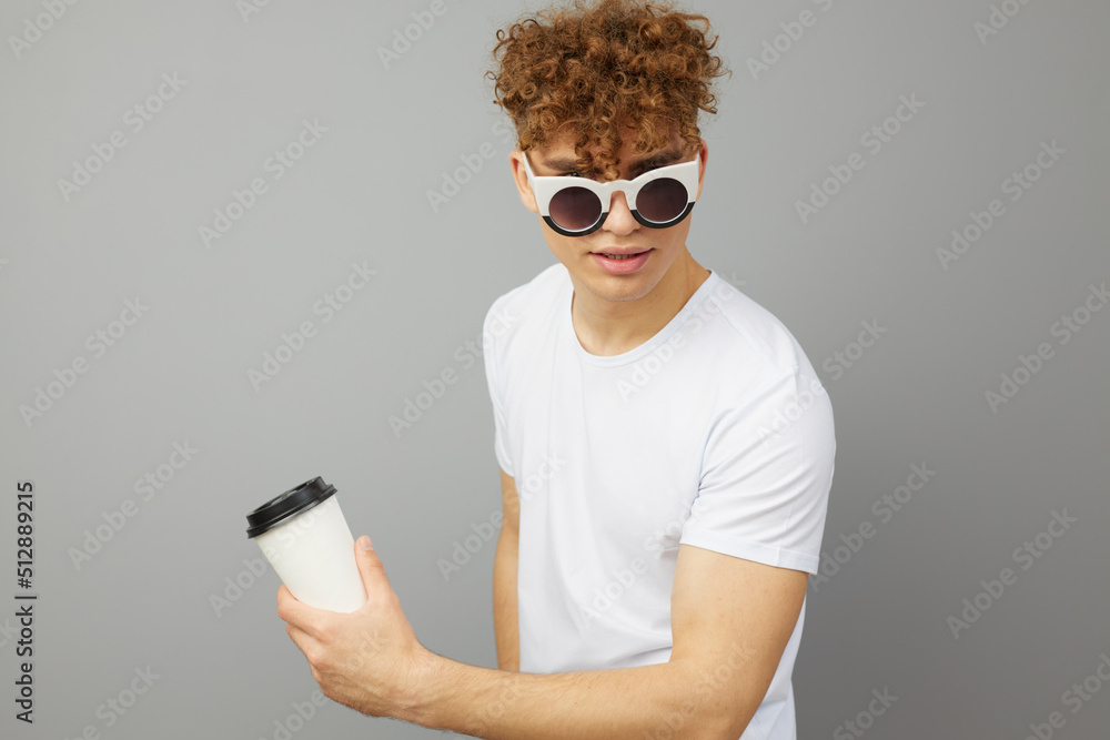 a young man of athletic build stands dressed in a white T-shirt, with sunglasses on his face and holding a white glass in his hand looking at the camera, standing sideways a young man of athletic build stands dressed in a white T-shirt, with sunglasses on his face and holding a white glass in his hand looking at the camera, standing sideways