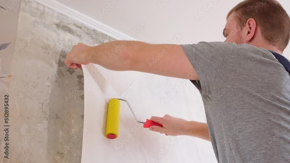 A man rolls out a canvas of white wallpaper with a suture wallpaper yellow roller. Removing air