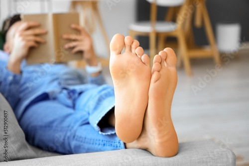 Fototapeta Young barefoot man reading book on sofa at home, closeup
