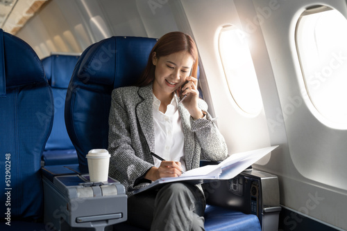 Business young asian woman talking on phone during working for paperwork financial report on airplane, flight in plane photo