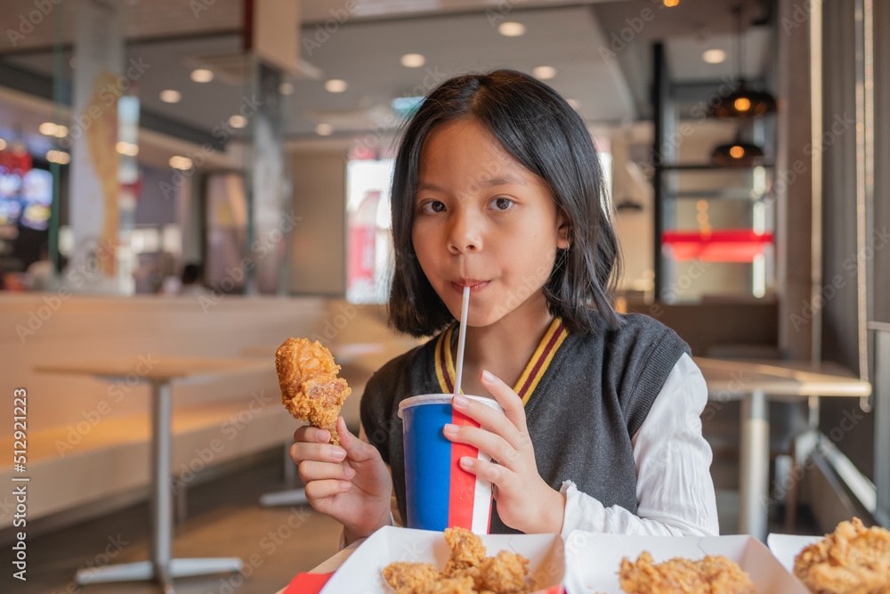 Close up portrait of a satisfied pretty little asian girl eating fried chicken and french fries, sparkling water in the restaurant. Unhealthy food concept, close up Close up portrait of a satisfied pretty little asian girl eating fried chicken and french fries, sparkling water in the restaurant. Unhealthy food concept, close up