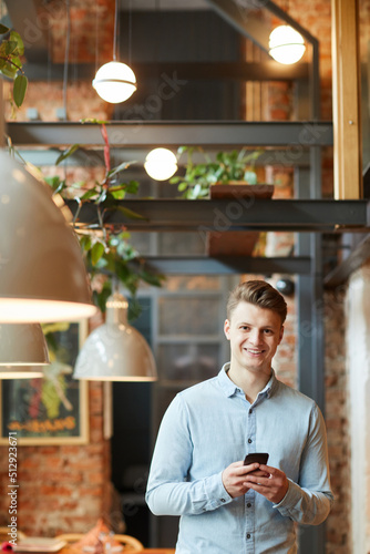 Portrait of positive handsome young tester in light blue shirt standing in loft coffee shop and using smartphone