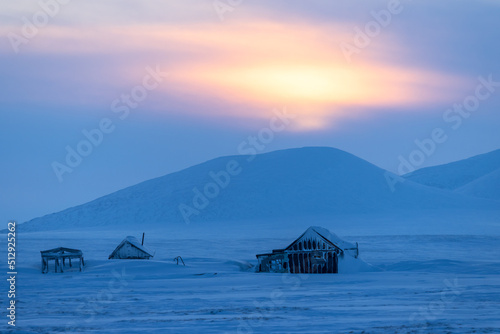 Winter Arctic landscape. Abandoned houses among the snow-covered tundra on the background of snow-capped mountains. Sunset over the mountain. Harsh polar climate. Siberia, The Far North of Russia.