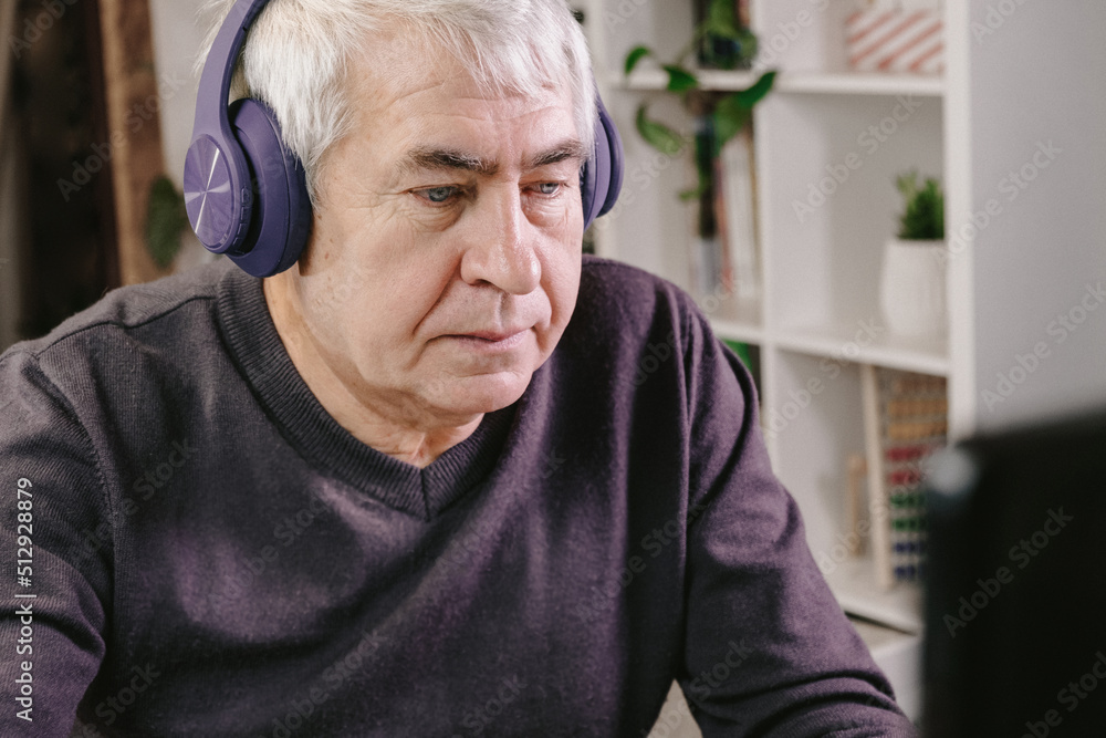 Cheerful older person with headphones sitting at table at cozy room and working on computer. Old Man using laptop, watching online program, funny video. Gray hair man received good positive news Cheerful older person with headphones sitting at table at cozy room and working on computer. Old Man using laptop, watching online program, funny video. Gray hair man received good positive news