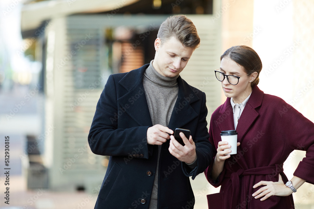 Pensive young business colleagues in stylish coats using new mobile app on move while testing it together Pensive young business colleagues in stylish coats using new mobile app on move while testing it together
