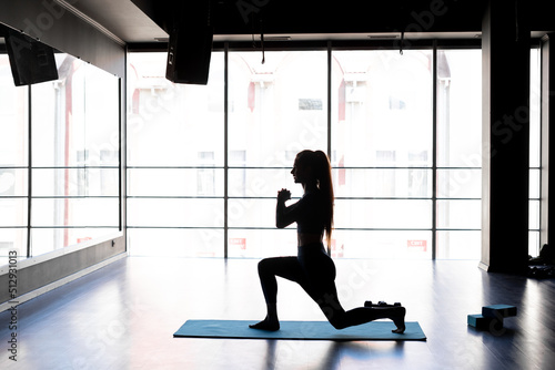 Attractive young girl is exercising in the gym near the window, silhouette of a woman doing fitness using a mat for stretching legs indoors. Sport, lifestyle and health