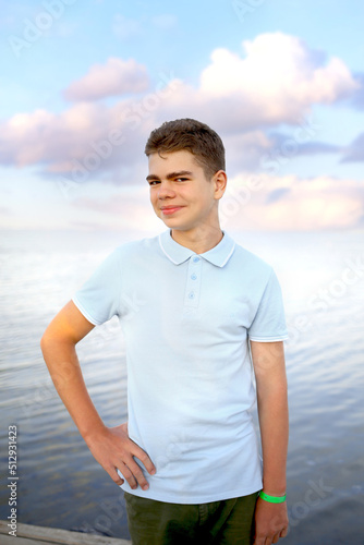 Cute teen boy in a blue t-shirt and green shorts looking at the camera with a smile. Background of the sunset sky on the sea. Childhood. Holidays and rest. Joy concept