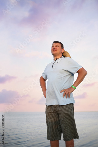 A young man in a blue T-shirt and green shorts standing on the pier. Stunning sunset sky background on the sea. Travel and recreation. Tourism concept