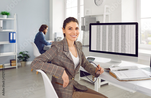 Portrait of professional financial accountant at work. Happy cheerful beautiful young woman in suit sitting at office desk with modern desktop computer, holding glasses, looking at camera and smiling