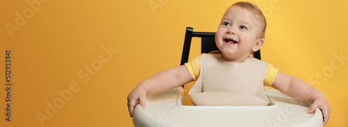 Cute little baby wearing bib in highchair on yellow background, space for text. Banner design photo